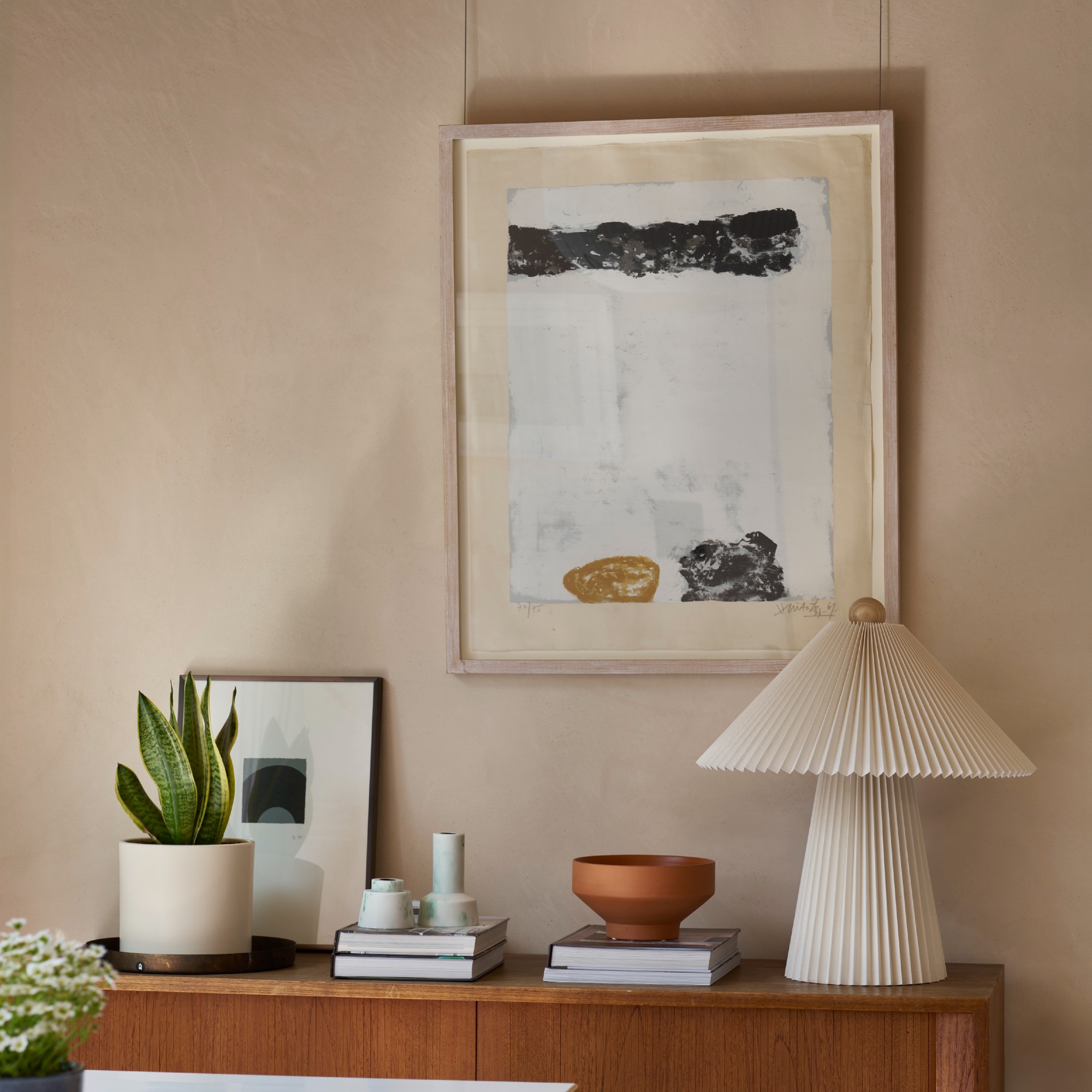 A light peach-painted wall with an abstract artwork and a sideboard placed against it displaying decorative items and a pleated paper table lamp