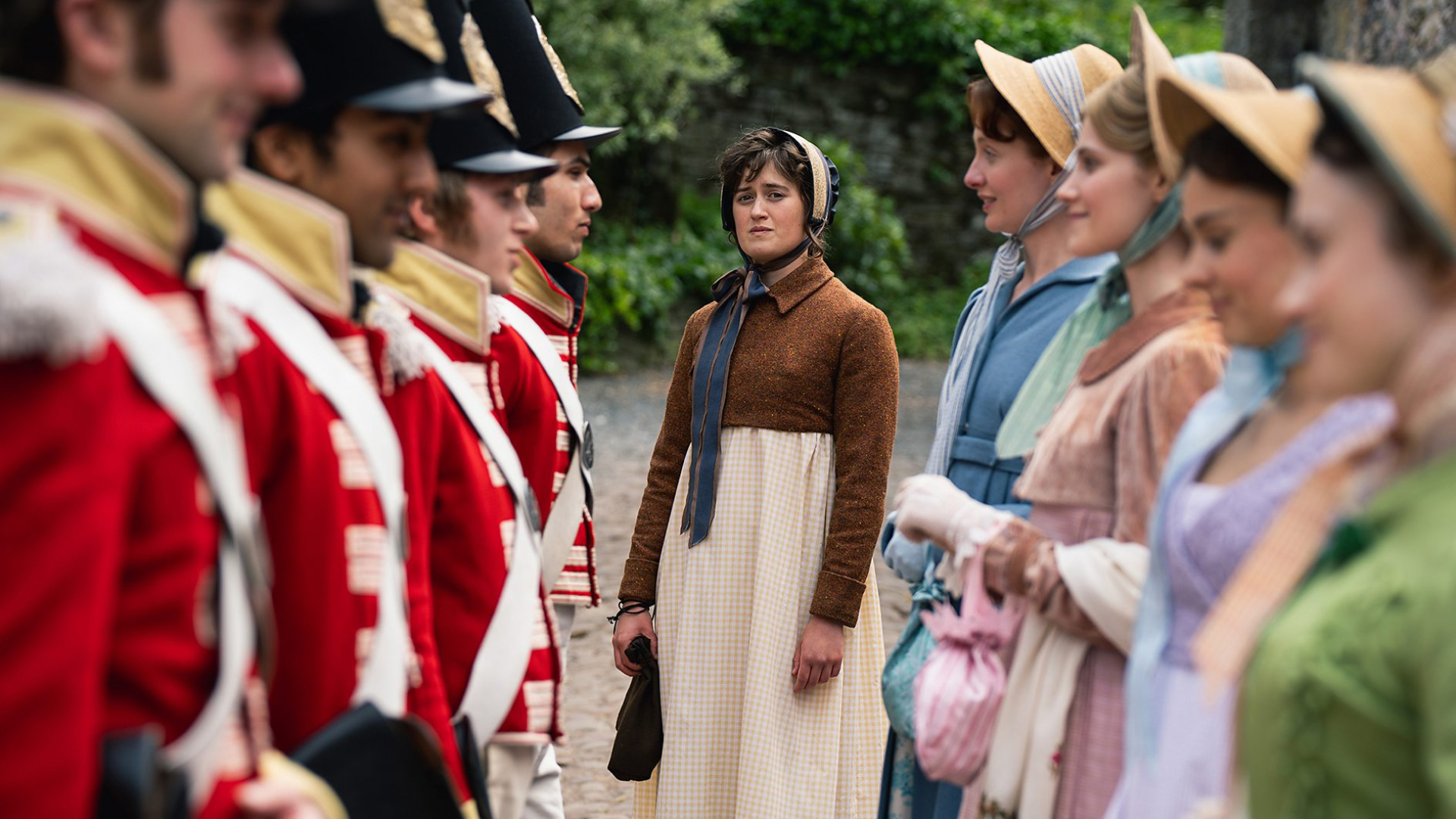 A Regency-era woman (Ella Bruccoleri as Mary Bennet) stands alone while framed by a row of red-coated soldiers and another row of Regency women, in BritBox's 'The Other Bennet Sister.'