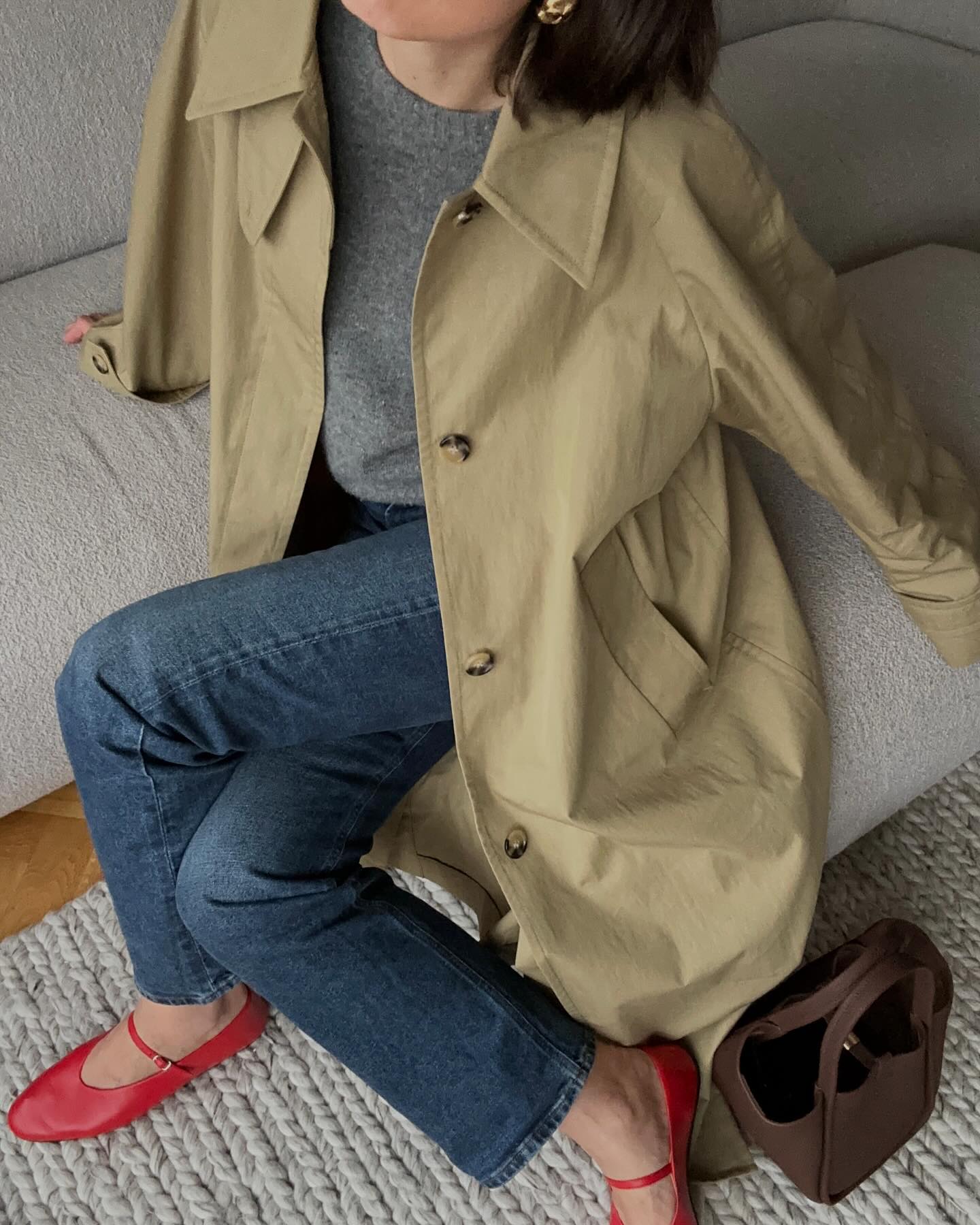 a female fashion influencer poses on a gray sofa wearing an effortless outfit with a trench coat, gray sweater, straight-leg jeans, and red Mary Jane flats
