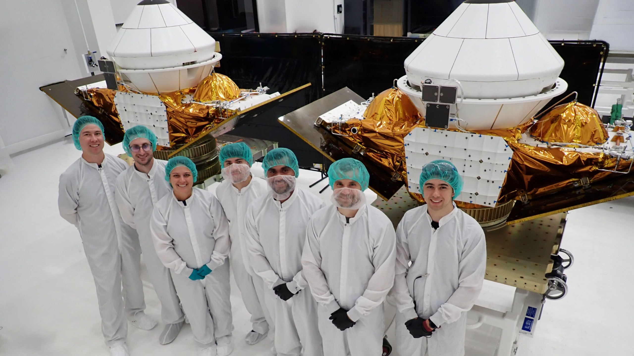 seven people in white lab coats stand in front of two white cone-shaped spacecraft inside a white-walled cleanroom