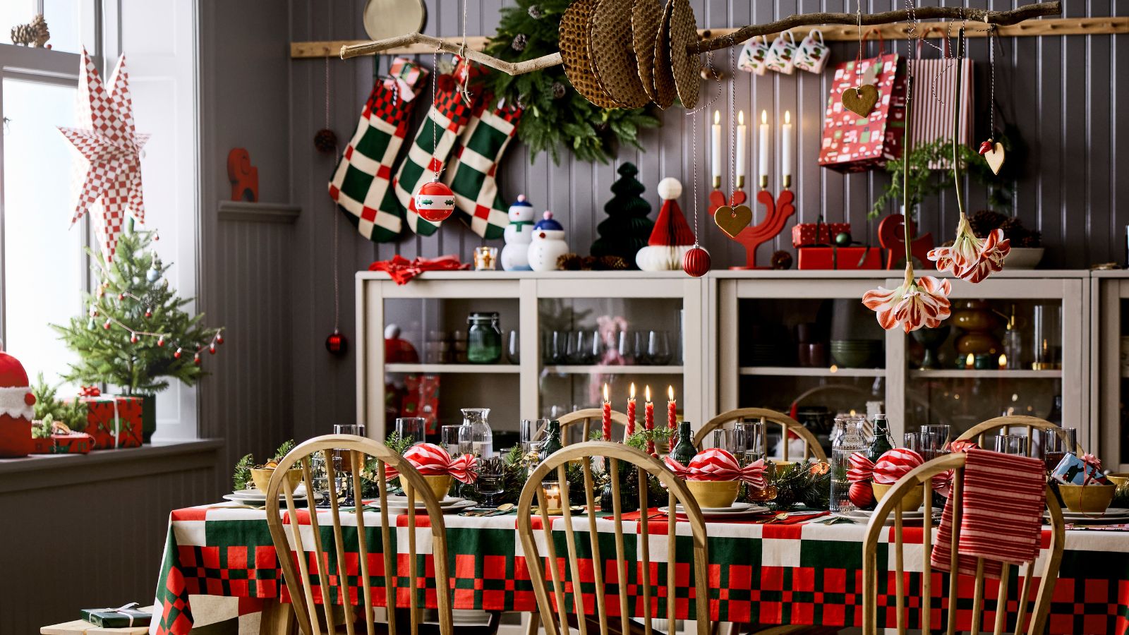 A dark grey panelled dining room styled for christmas with a large table with wooden chairs, styled with red and green christmas tablecloth and decor