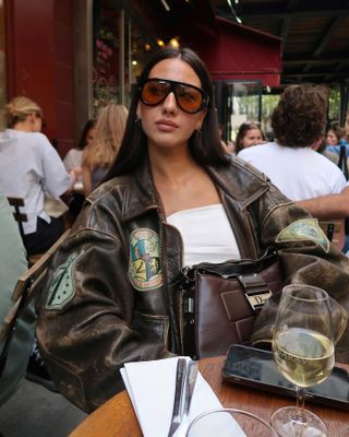 Italian woman wearing leather jacket, white tube top, and oversize Tom Ford sunglasses while sitting at a table.