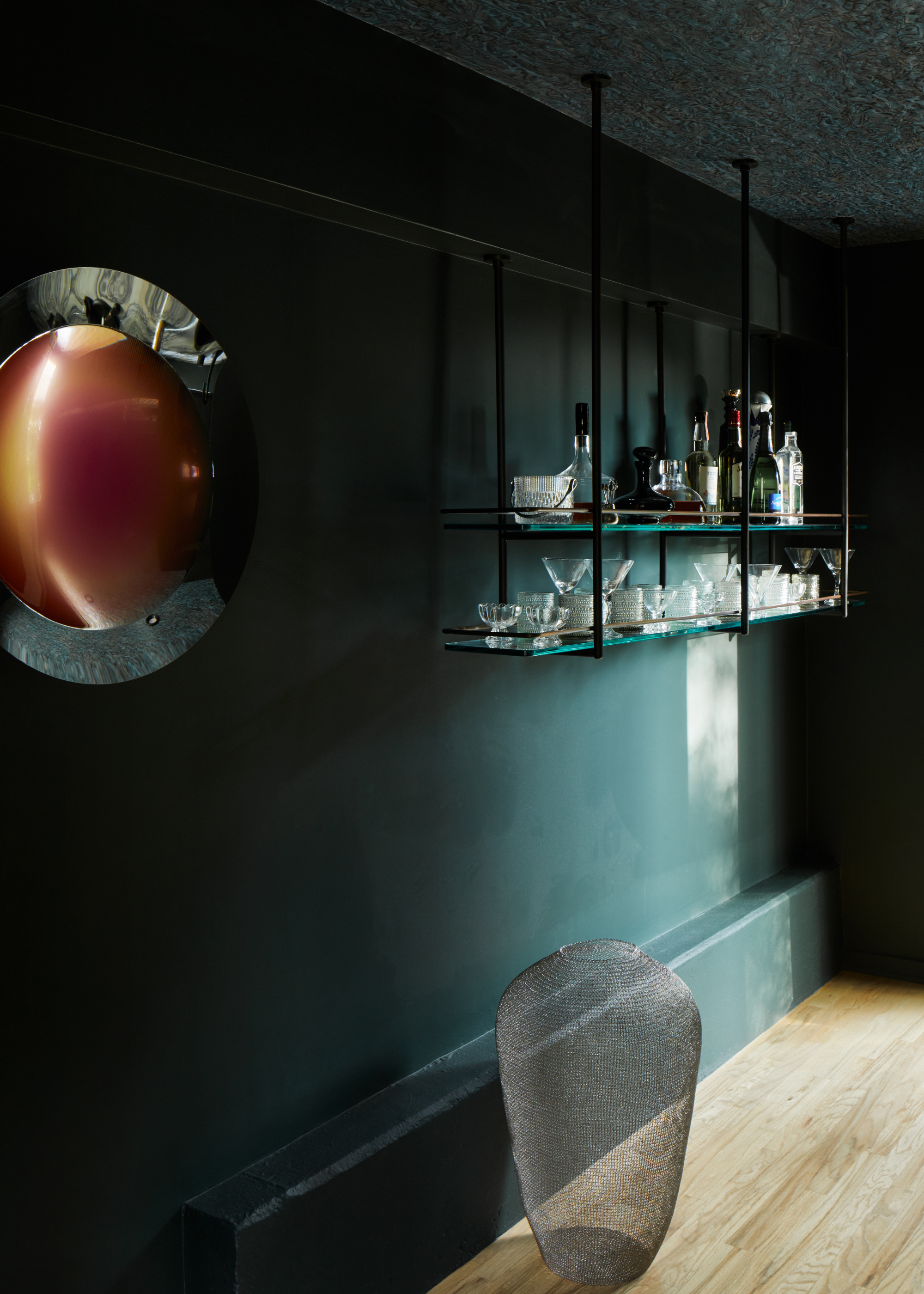 A dark green bar room, with a floating rack of spirits and glassware, a reflective wall hanging, a woven mesh vase and wood flooring