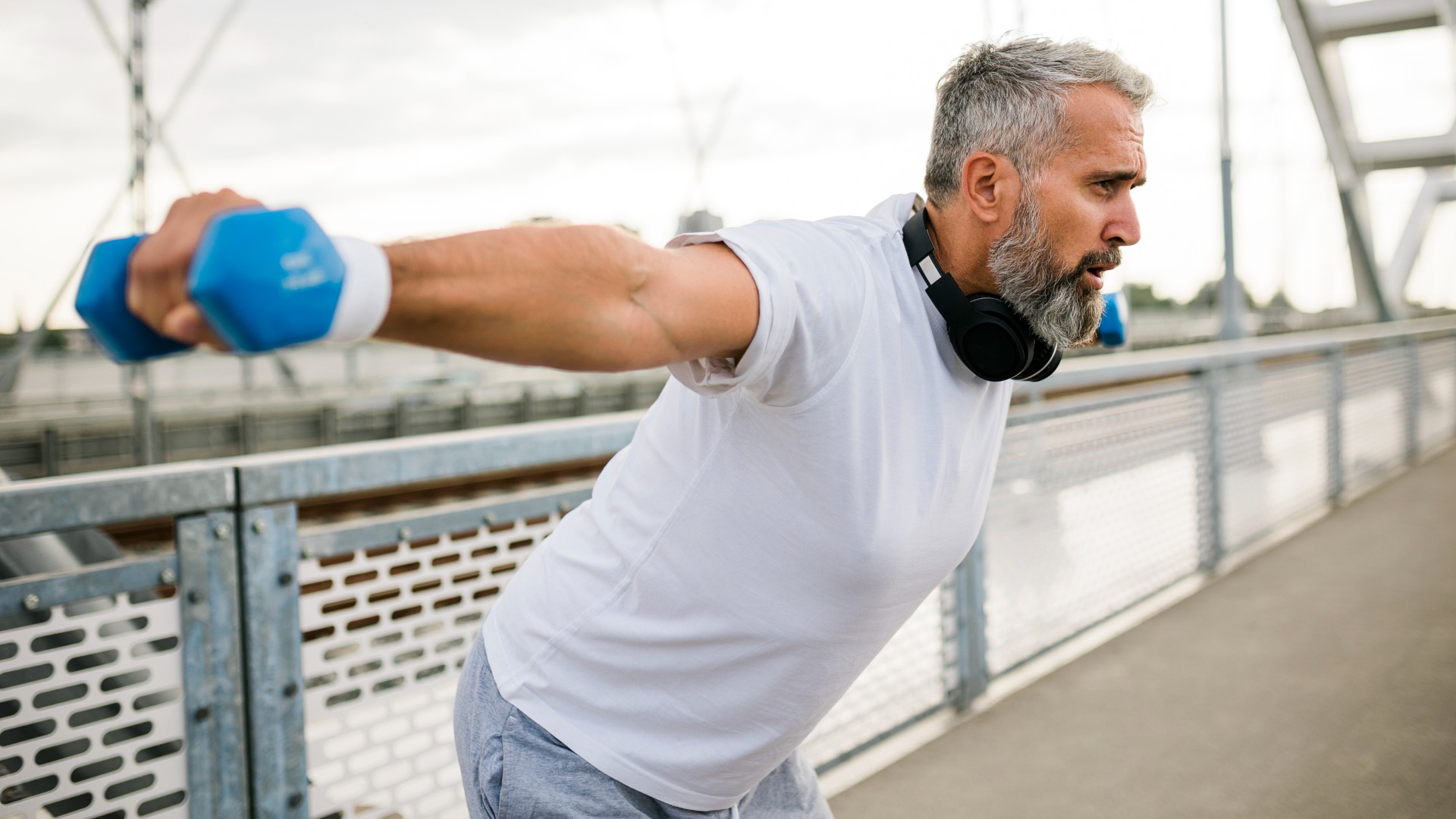Man with greying hair outside on a bridge performing shoulder flyes using light dumbbells