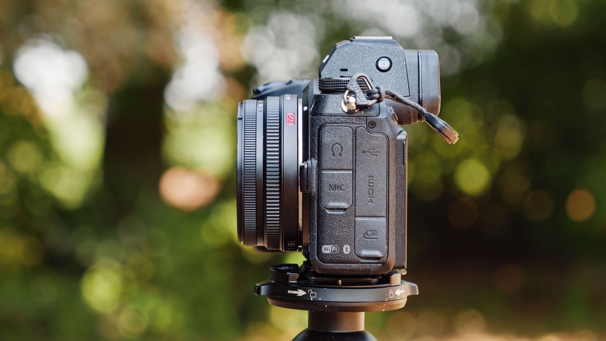 Side profile of the Viltrox AF 26mm F2.8 EVO 'pancake' lens attached to a Nikon mirrorless camera, there's dappled light through a tree behind it