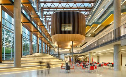 Interior of the Vancouver’s University of British Columbia. Metal tables with black and red chairs are arranged throughout the space. Large concrete columns support the level above. Wide stairs are to the left, that lead to the outside.