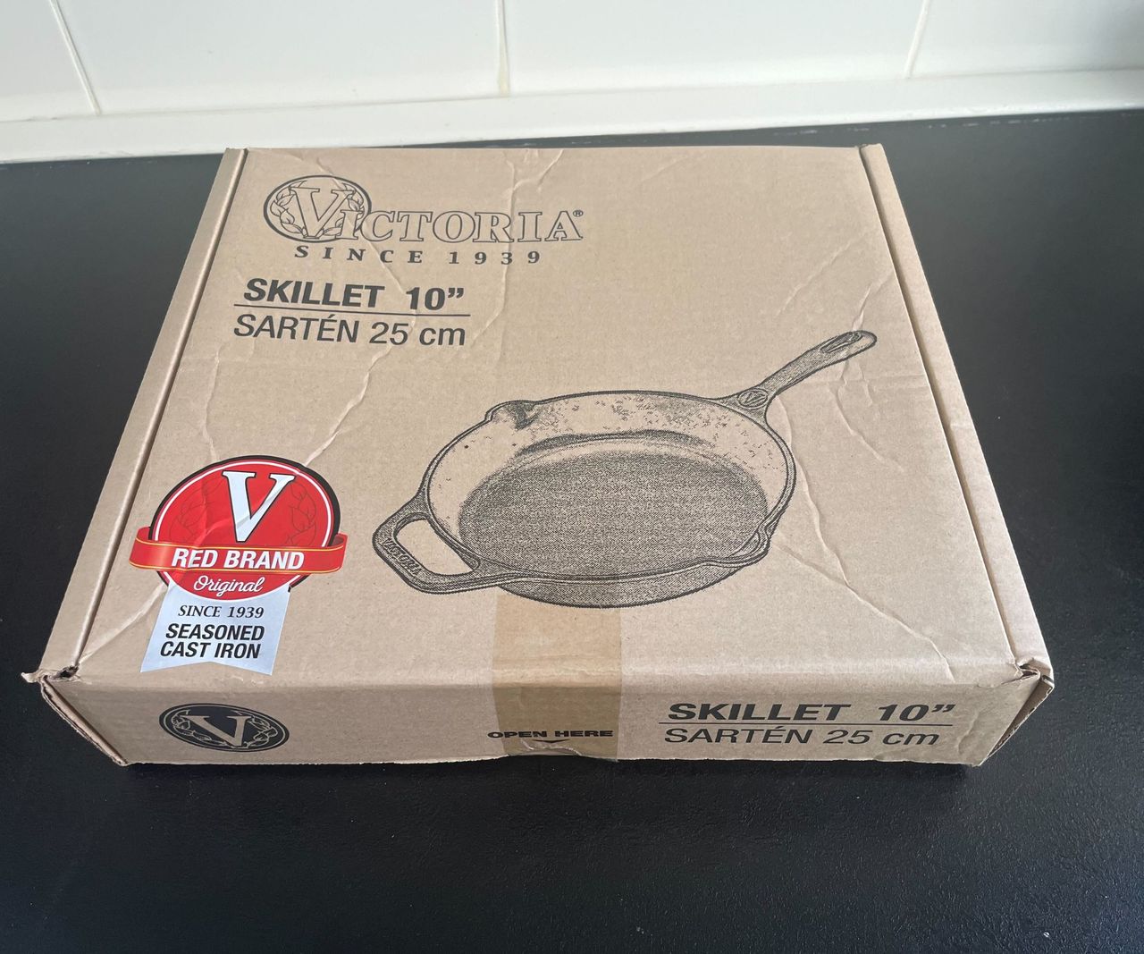 A brown cardboard box with a Victoria Cast Iron Skillet printed on the front on a black kitchen counter.
