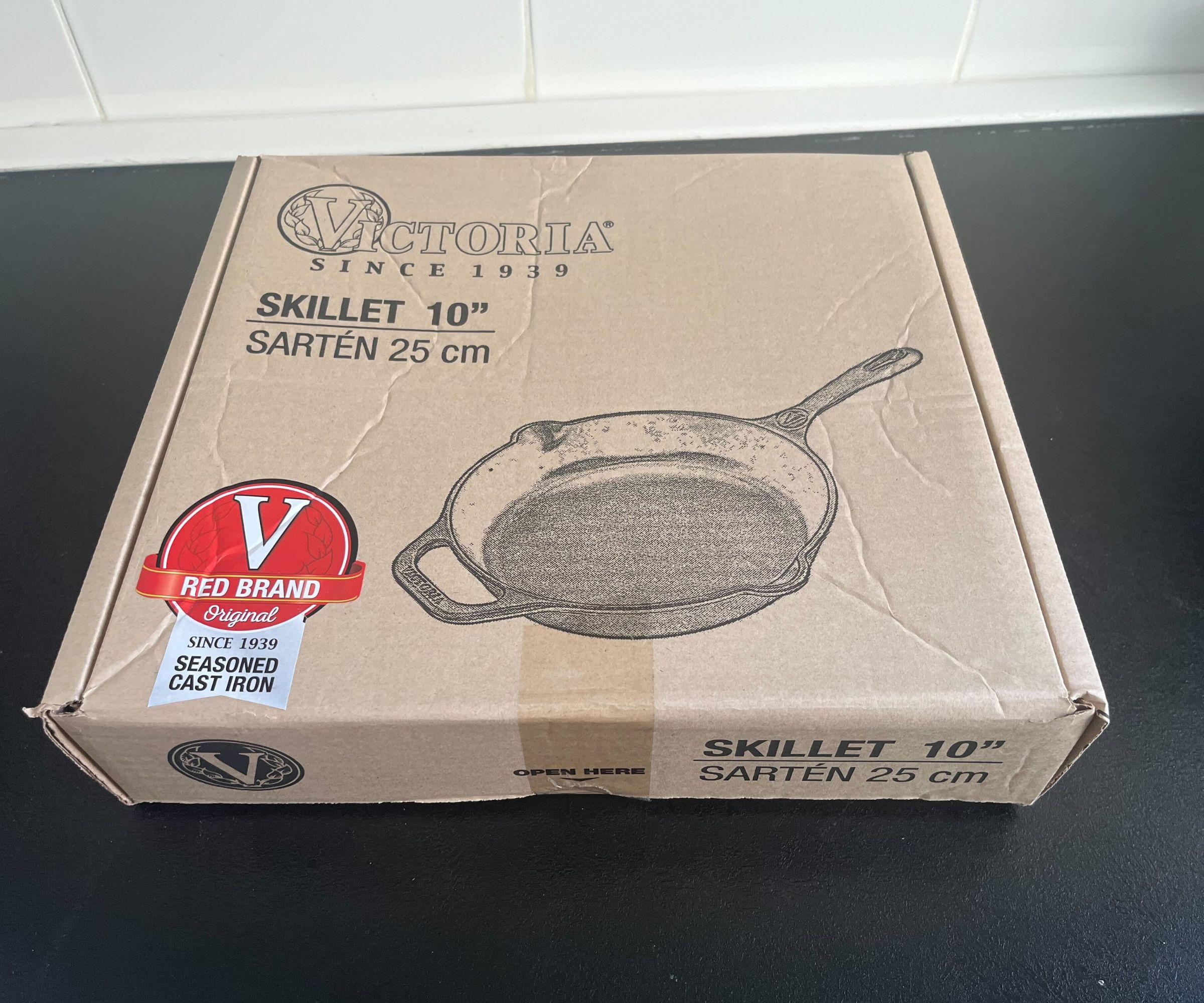 A brown cardboard box with a Victoria Cast Iron Skillet printed on the front on a black kitchen counter. 