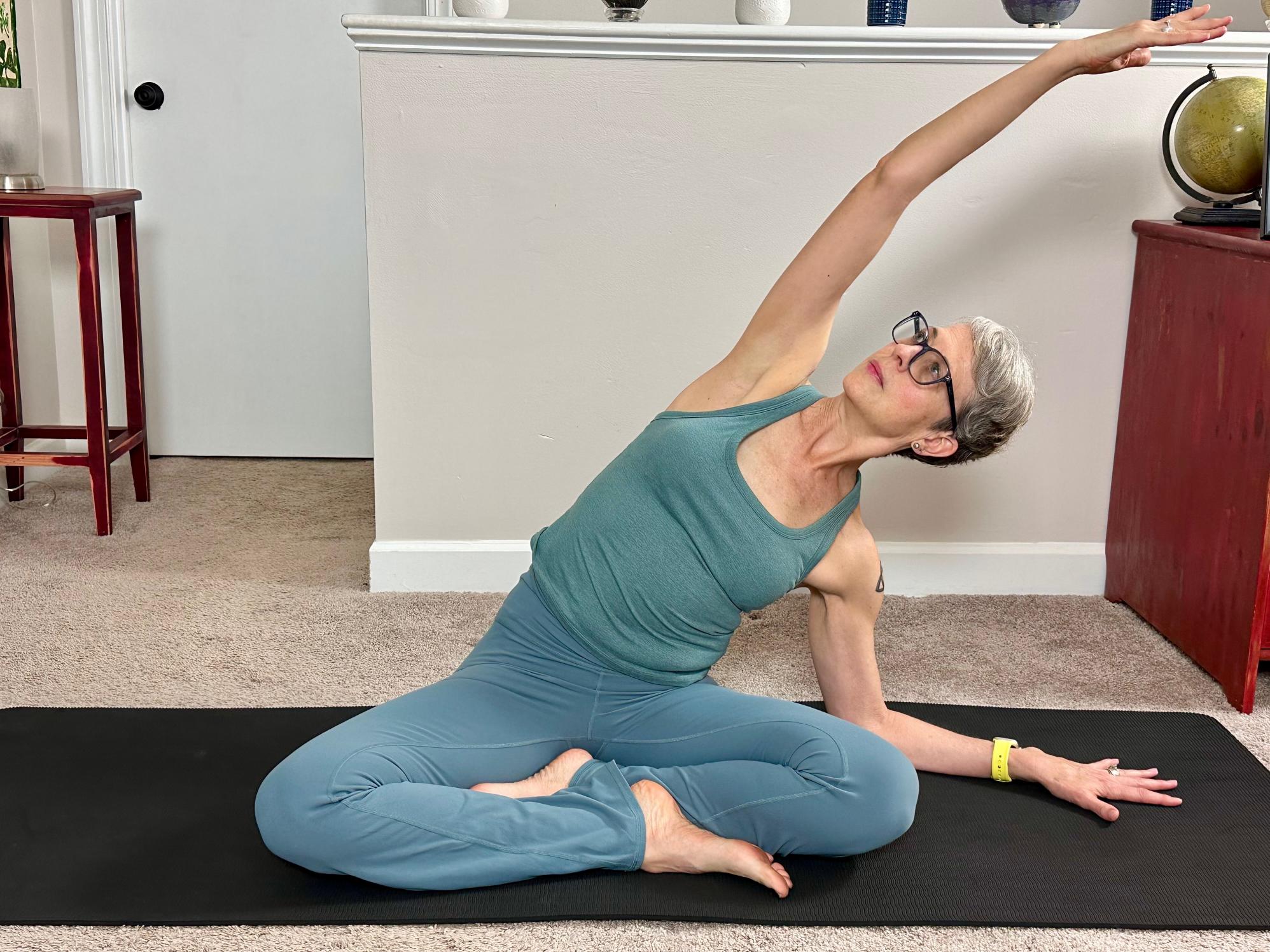 woman sat cross legged reaching to one side for a side body stretch on a black yoga mat in a living room