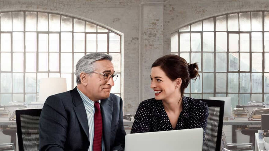 A still from the movie The Intern in which Robert DeNiro and Anne Hathaway play Ben and Jules and they’re sat in an office smiling at each other.