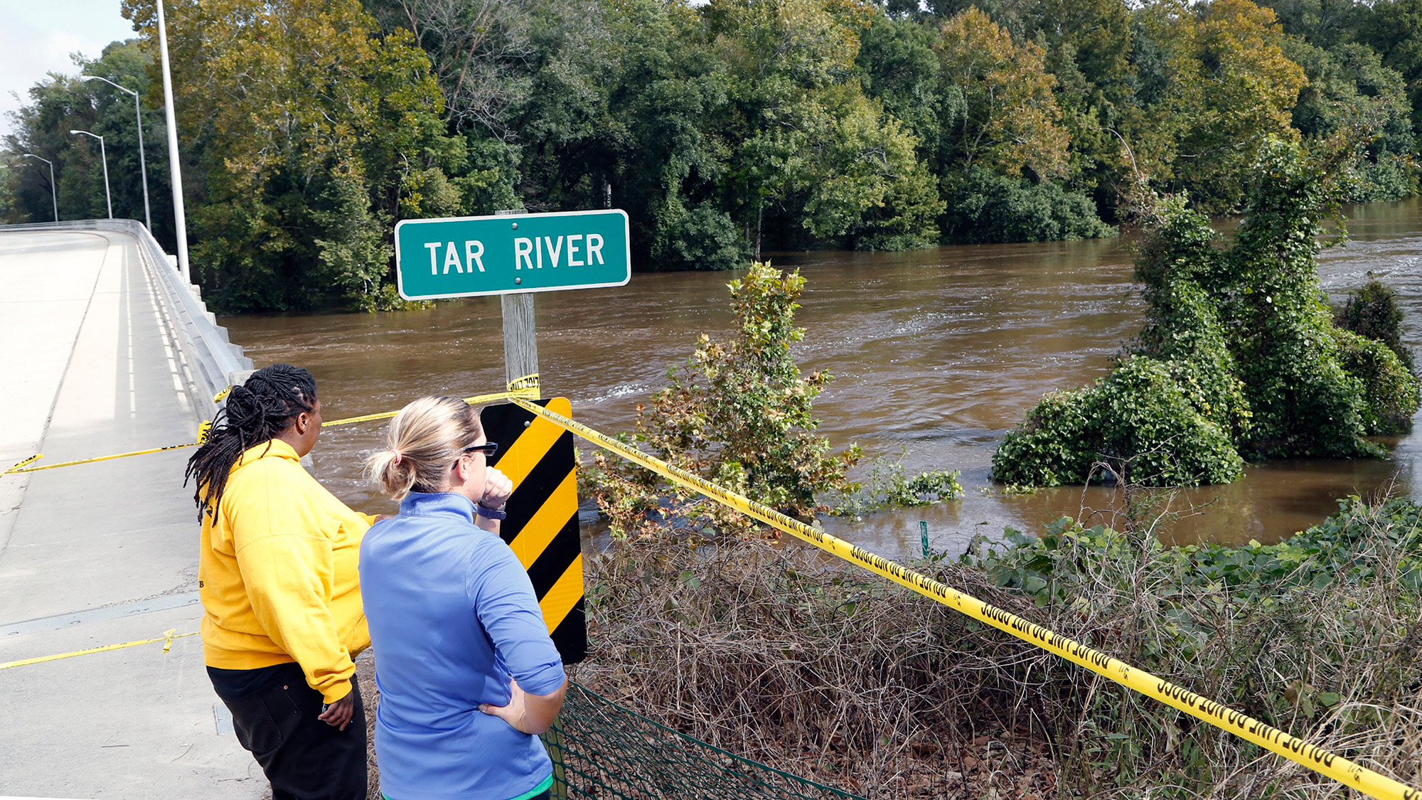 Two women stand next to a river with the road sign "Tar River" next to them, with the banks of the river overflowing.