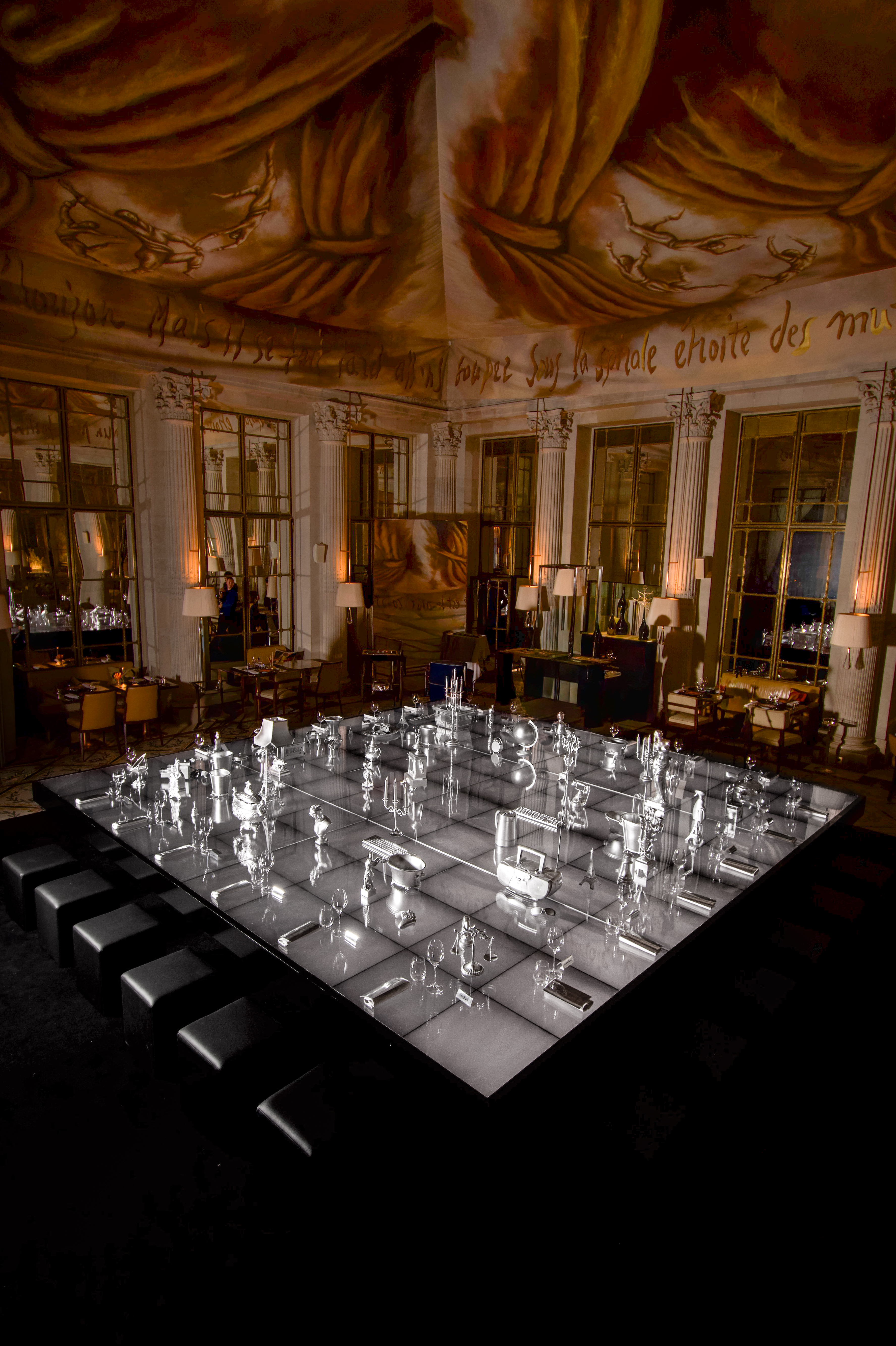 A monochromatic installation set inside a palatial hotel with mural ceilings, featuring a back-lit white surface topped by lots of silver objects, around which a man with dark long hair poses.