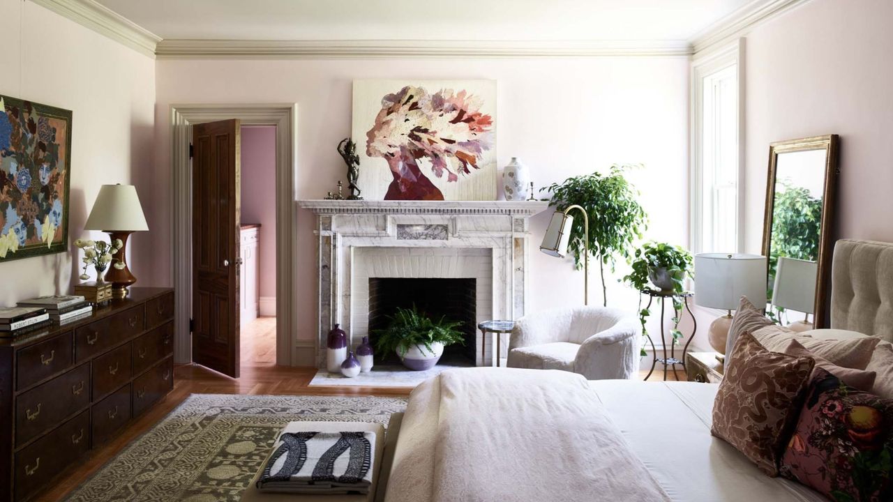 bedroom with original marble fireplace with artwork above it, plants on stands by the window, a wooden dresser along one wall, a patterned rug, and a bed with lots of throw pillows