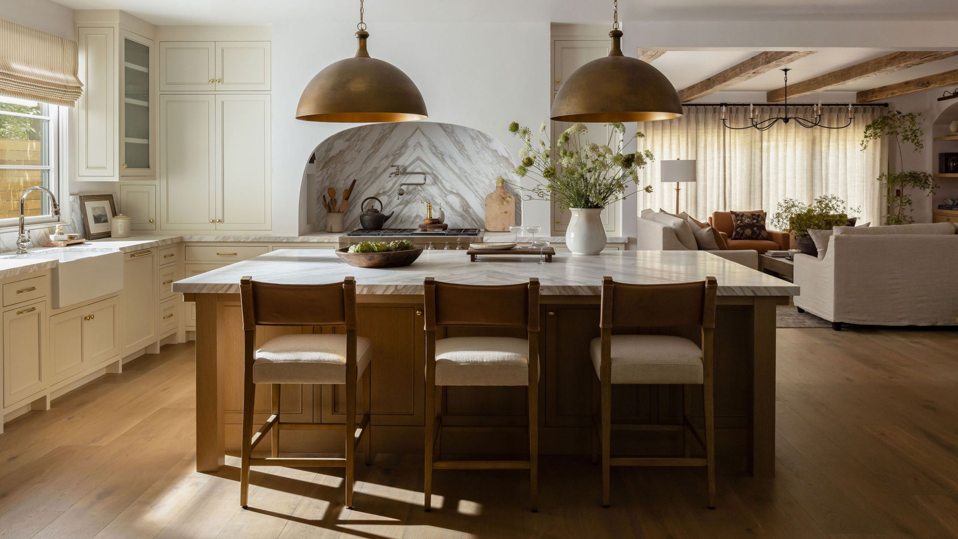 a neutral farmhouse style kitchen with off white cabinetry, arched cooker hood, oak kitchen island with wooden stools and large brass pendants above