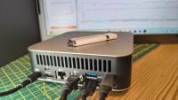 A Geekom A7 Max mini PC with multiple cables connected sits on a desk, with a USB flash drive on top and a computer monitor displaying graphs in the background.