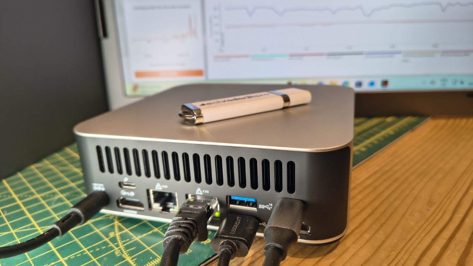 A Geekom A7 Max mini PC with multiple cables connected sits on a desk, with a USB flash drive on top and a computer monitor displaying graphs in the background.
