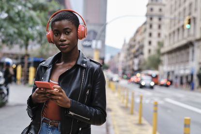 Woman wearing headphones and jacket using smart phone while standing in city