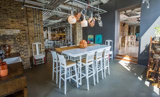 The Grain Store, London, UK. A large white and brown table with white high chairs around it and lights, pots and pans hanging above it.
