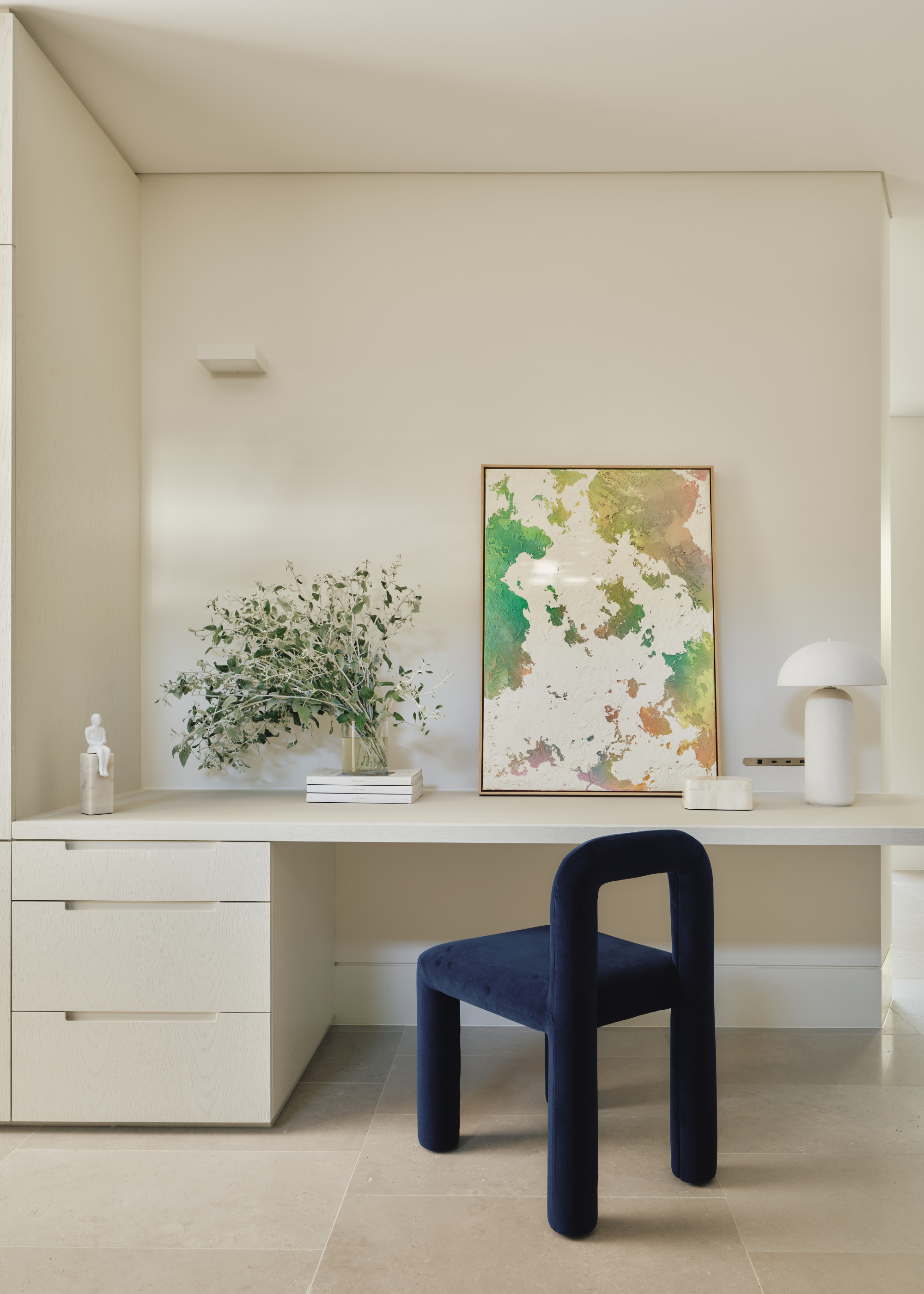 A minimalist study space with a floating desk, a leaning painting, a white table lamp, a stack of books, and a dark blue chair
