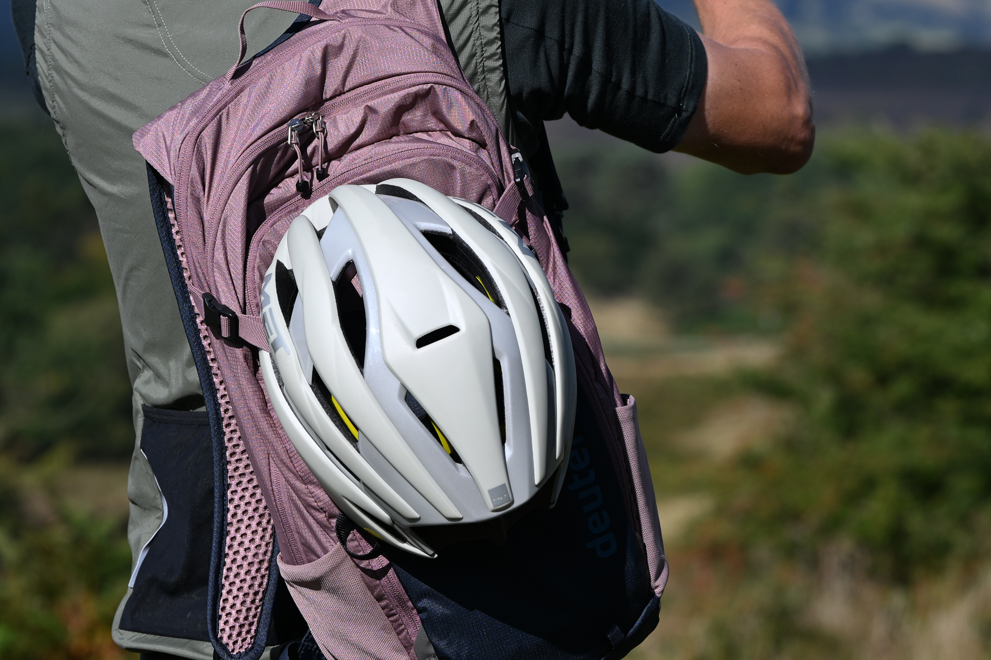 close up of a man wearing a green jersey, black shorts and a green gilet with a pink and blue rucksack, with a helmet attached to the backpack