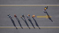 An overhead shot of five people walking along a road towards a yellow arrow pointed in their direction of travel, with a sixth person splitting off from the group to make their own way towards the top of the frame.