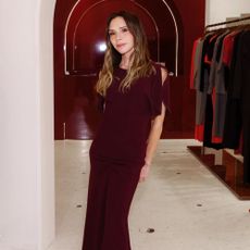 Victoria Beckham stands inside her stop wearing a burgundy dress with cream heels.