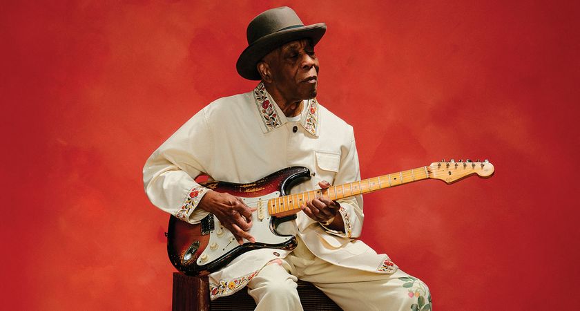 Buddy Guy wears a matching cream jacket and pants with floral stitching and a wide-brimmed hat as he sits and plays a Fender Stratocaster against a deep red background.