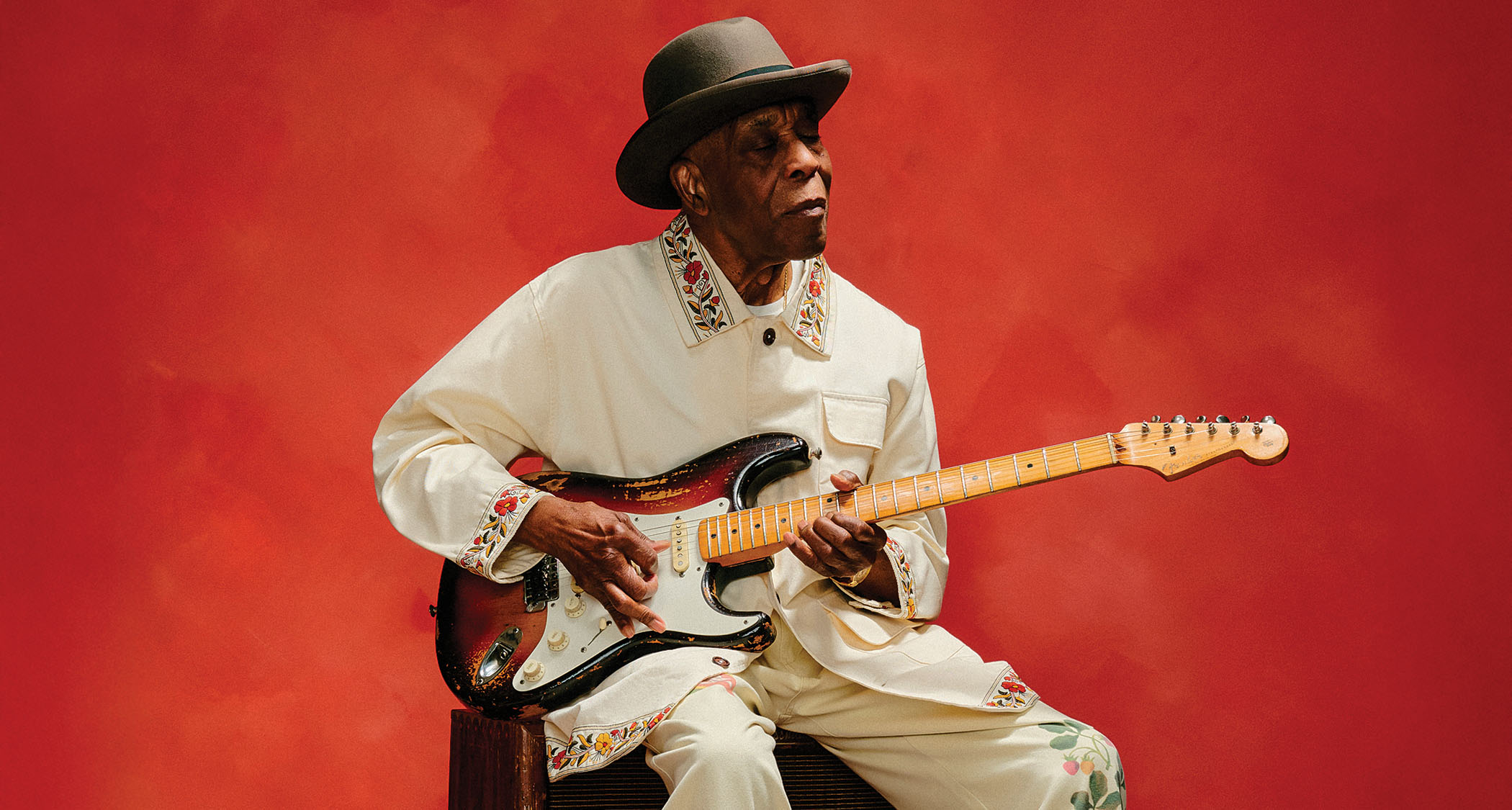 Buddy Guy wears a matching cream jacket and pants with floral stitching and a wide-brimmed hat as he sits and plays a Fender Stratocaster against a deep red background.
