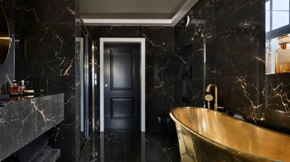 An all black marble bathroom with a statement gold freestanding bathtub to the right
