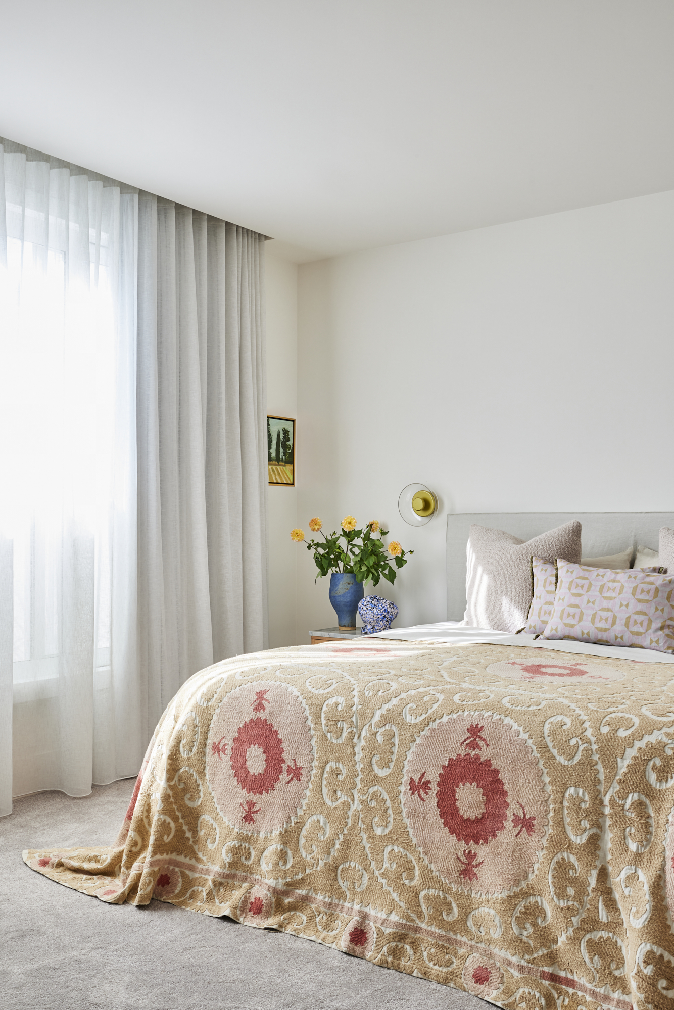 white bedroom with sheer curtains, a gray bedhead and a patterned blanket. on the bedside table is a vase with flowers and a wall sconce above