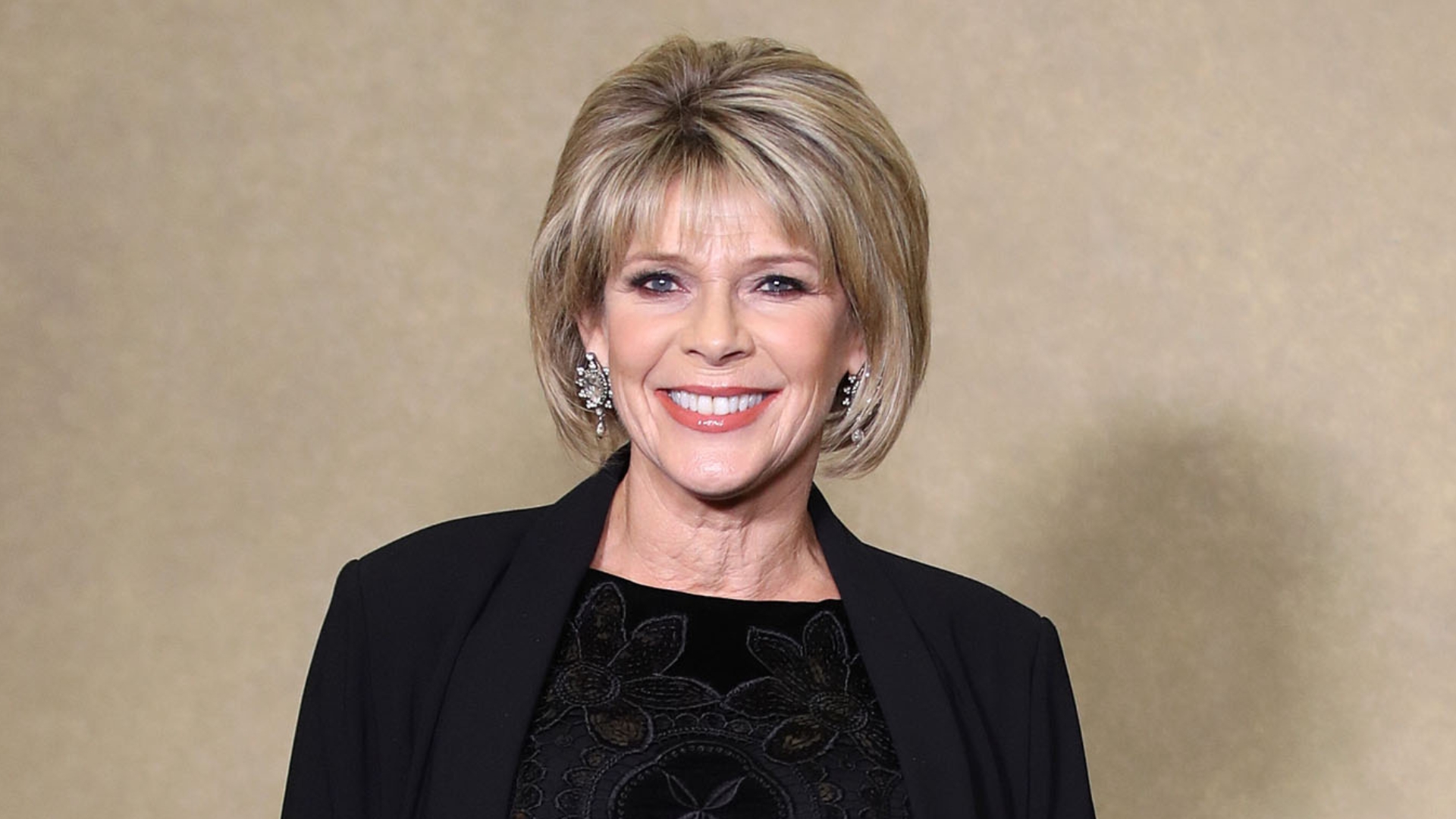 Ruth Langsford smiling with blonde bob and fringe, wearing crystal earrings, black patterned top and black blazer