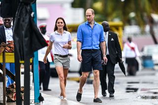 Kate Middleton wearing a white polo and shorts walking on a rainy road with Prince William, dressed in a blue shirt and navy shorts