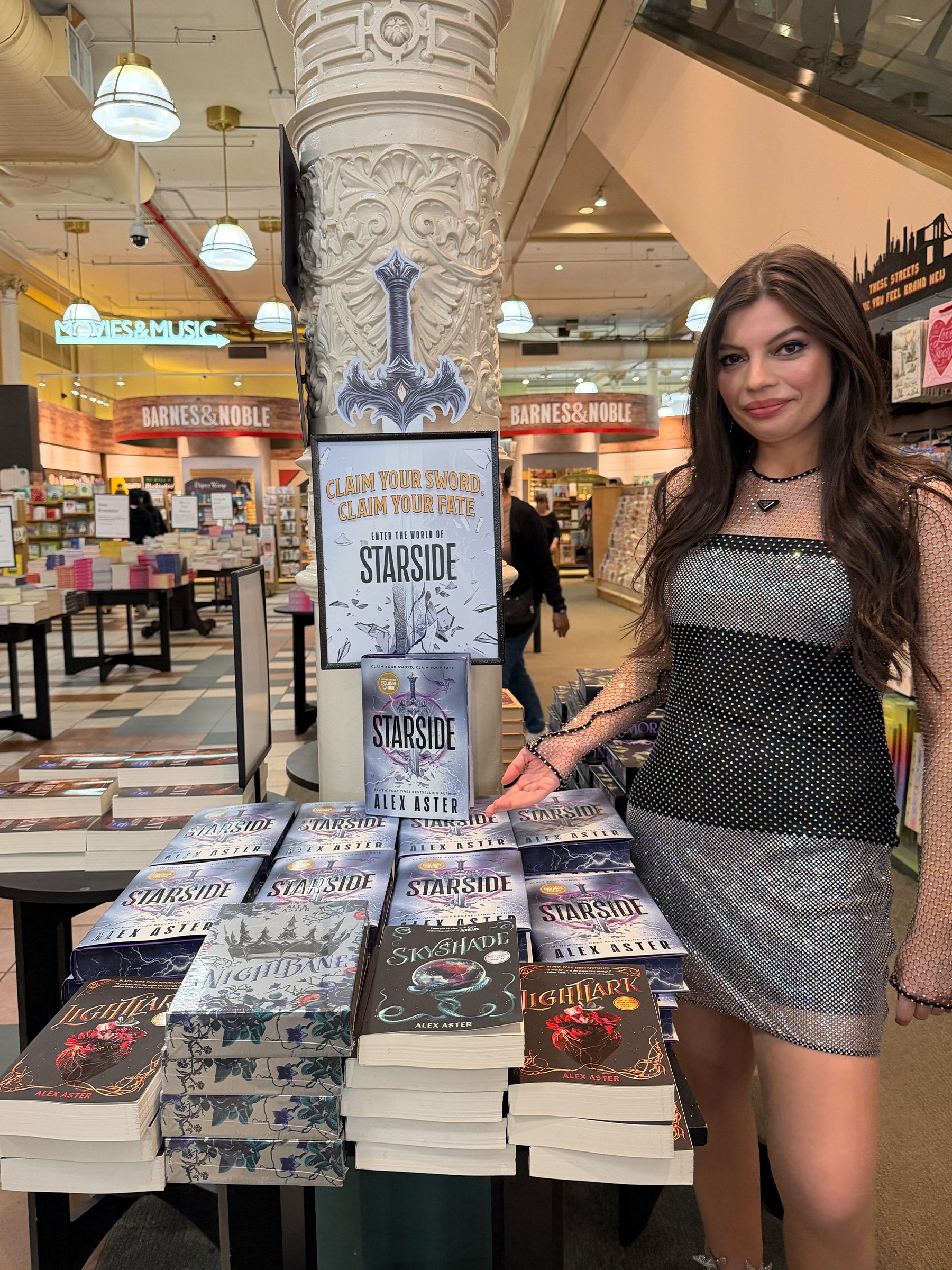 Alex Aster poses near her Starside book in a book store wearing a silver and black Prada sequin chainmail dress.