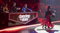 The Dancing with the Stars judges stand behind the table as they watch dancers in front of them.