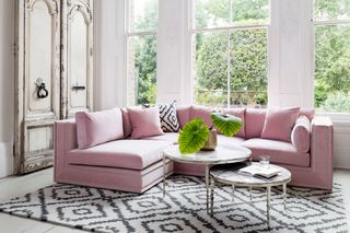 A pastel pink L-shaped couch for a small living room with large windows, a monochrome geometric print rug and a white color scheme.