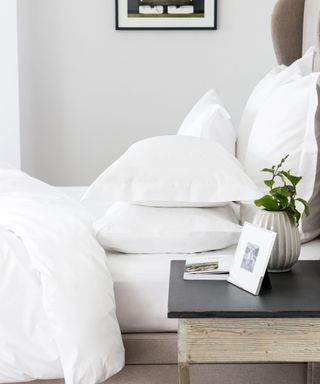 A side view of a white bed with a duvet and pillows, with a gray wall beyond it and a black and gray wood nightstand with a photo frame and plant next to it