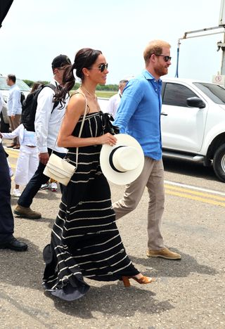 Meghan Markle wearing a black maxi dress and carrrying a straw bag and hat walking outside next to Prince Harry