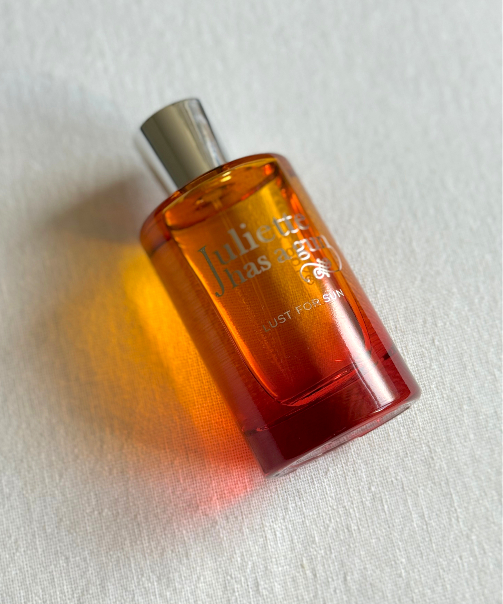 A bottle of Juliette Has A Gun Lust For Sun laying at an angle against an off-white linen background