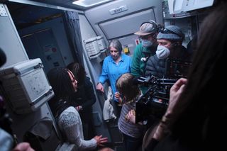 A group of people gathered inside an airplane being filmed by a large camera