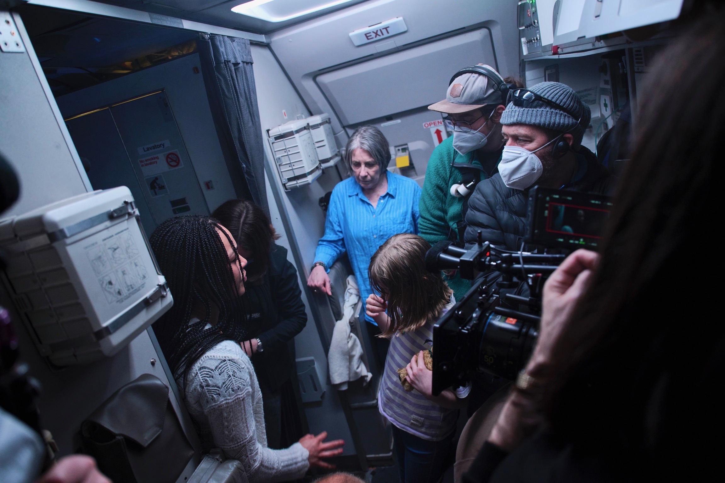 A group of people gathered inside an airplane being filmed by a large camera