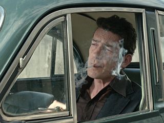 Image a still from the film 'The Secret Agent', a man in a car smoking a cigarette