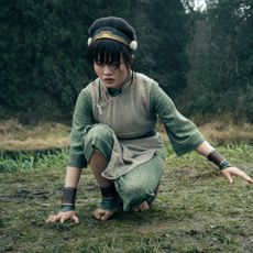 A girl (Miya Cech as Toph) kneels while touching the grass in a forest clearing, in a still from 'Avatar: The Last Airbender' season 2.