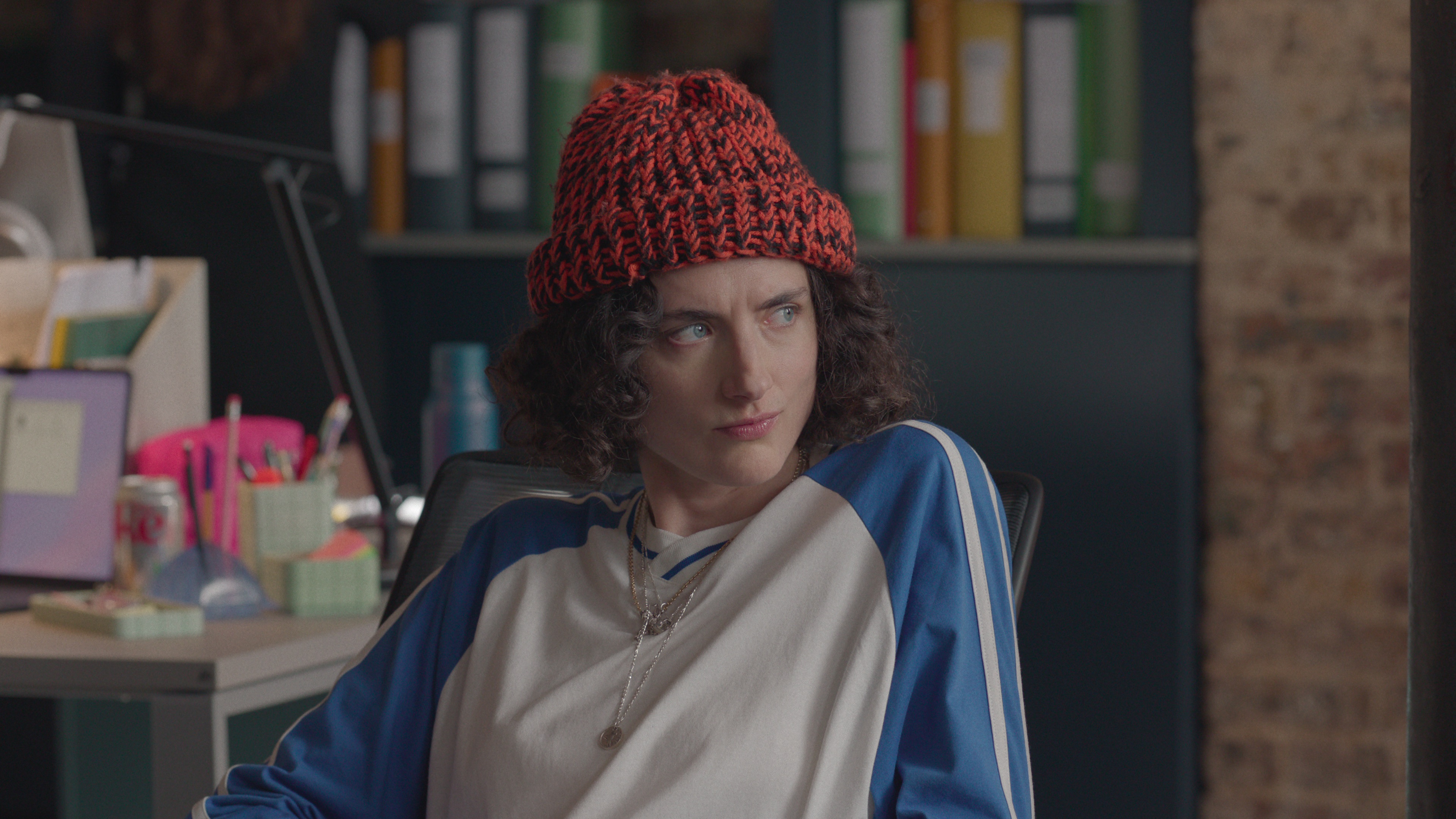 daisy bevan as josie wearing a beanie sitting at her desk in the office in too much