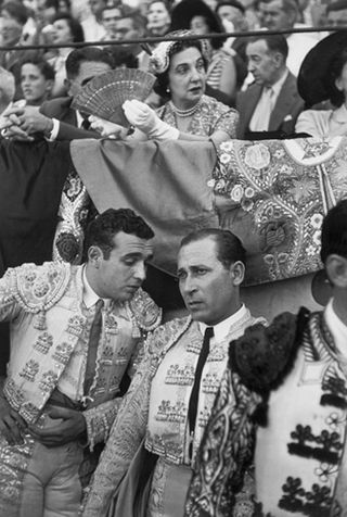 Black and white image of a crowd San Fermines, Pamplona, Spain. 1952, older lady smartly dressed with a spanish fan, matadors are at the bottom of the shot two are deep in conversation