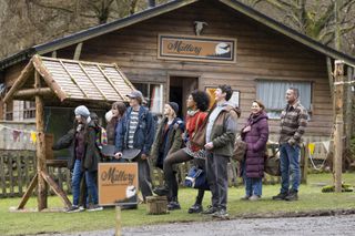 Dani (Kara Tointon) and Jimmy (Will Mellor) stand outside a log cabin with a sign saying The Mallory Outdoor Activity Centre, with a group of pupils standing in front of them. Everyone is smiling and laughing, apart from a few of the pupils who look slightly bored