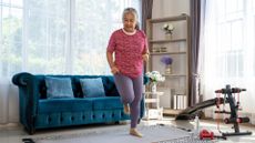 older woman standing in a living room setting moving onto one leg mid workout. there's some gym equipment to the side of her and a blue sofa and large window behind her.