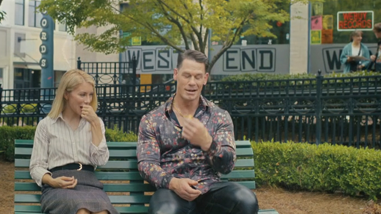 Jennifer Holland and John Cena sitting on bench in front of a store called West End Wax in Peacemaker