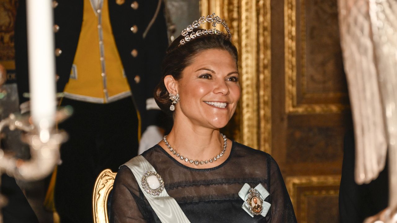 Crown Princess Victoria sitting at a table wearing a navy gown and sash with a diamond tiara 