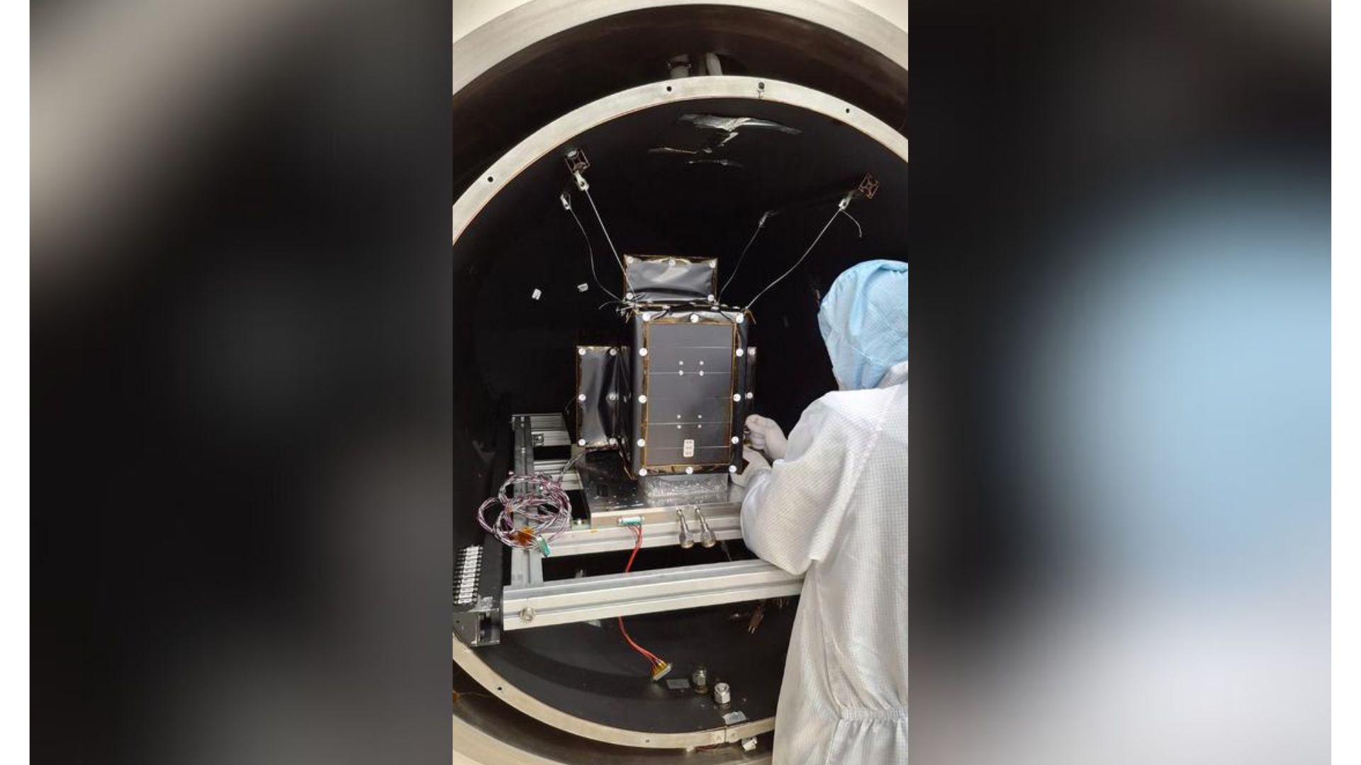 A person wearing a lab coat and hair net works on a box-shaped satellite in an experimental chamber