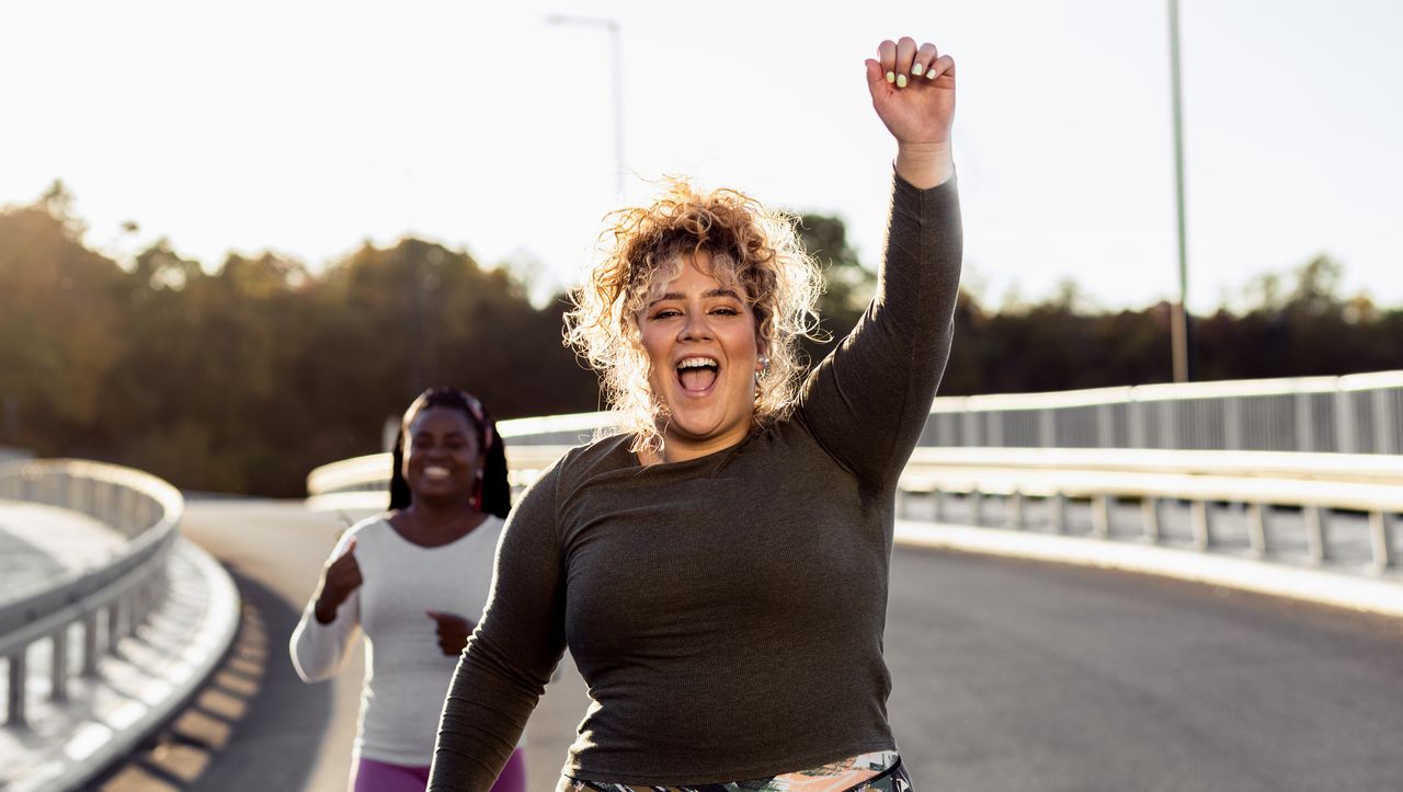 Woman runner on road with hand raised and mouth open in a joyful expression. She has curly hair and is wearing a long-sleeved black top. A smiling woman runner is in the background.